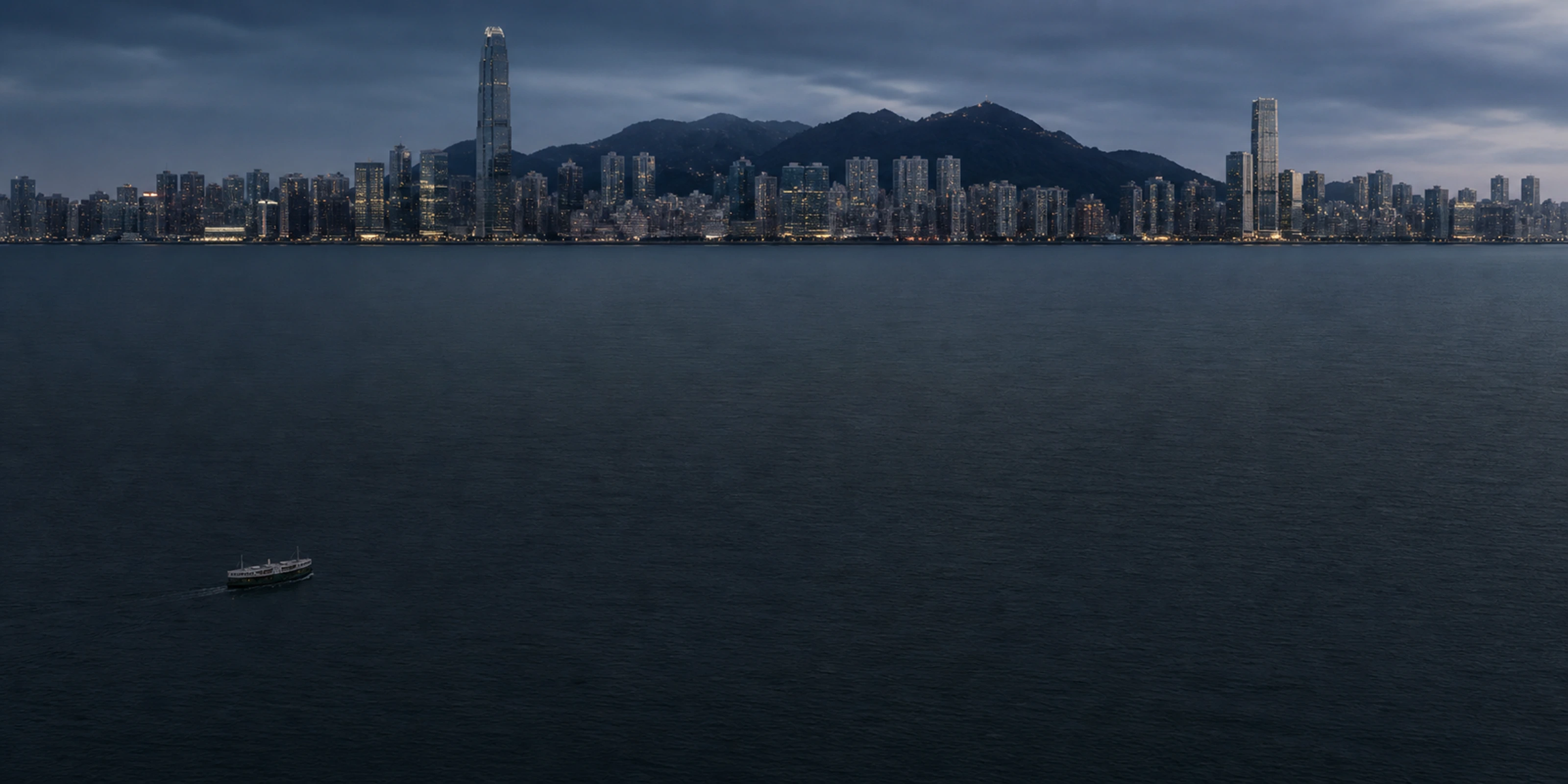 Victoria Harbour zur blauen Stunde: dunkles Wasser, die Skyline Hongkongs und Berge unter Wolken
